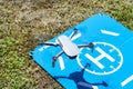 The drone hovers over the blue landing pad on the grass, the plastic propellers are on, and the camera on the gimbal is visible.