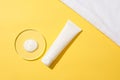 A drop of white cosmetics on a glass podium and an unlabeled cosmetic tube displayed on a yellow background. Build a cosmetic