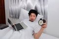 Drunk Asian man with laptop and alarm clock lying down on the bed and having hangover after party.