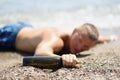 Drunk young man lying on seashore with bottle of wine in his hands closeup