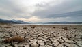 Dry cracked earth with sparse brown grass in foreground distant lake and mountain range under cloudy sky Royalty Free Stock Photo