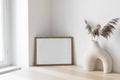 Dry grass, reed plant bouquet in modern ceramic vase. Empty photo frame mockup against white wall on wooden table near