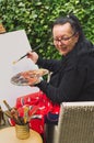 Old artist lady holding painter pallete, smiling and starting to paint on the empty canvas