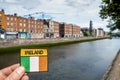 Dublin city center, Ireland - 07.06.2021: Badge with sign Ireland and Irish National flag in focus, Liffey river and old buildings Royalty Free Stock Photo