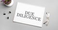 due diligence is written in a white notebook next to a pencil, black-framed glasses and a green plant