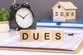 dues word in wooden blocks with small wooden house and alarm clock on gray background. symbolizes the timely payment of