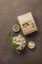 Dumplings in eco-friendly paper packaging for delivery and a bowl with dumplings, onions and sauces on a dark background