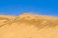 Dune against the background of a bright blue sky. The wind blows the sand off the ridge of the dune