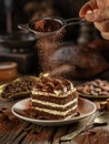 Dusting tiramisu-like cake with cocoa powder. Still life with slice of cake and coffee and cacao beans on wooden table Royalty Free Stock Photo