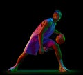 Dynamic image of teen boy in uniform, playing basketball against black studio background in neon light. Dribbling ball