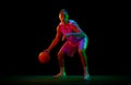 Dynamic image of teen boy in uniform, playing basketball against black studio background in neon light. Dribbling ball