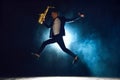 Dynamic shot of artistic man, solo performer jumping while playing saxophone on stage with backlights against dark Royalty Free Stock Photo