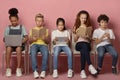 E-learning concept. Focused diverse schoolkids with gadgets and study materials sitting on chairs over pink background