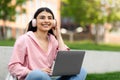 E-learning concept. Happy hispanic teen lady using laptop, wearing headphones listening to music, sitting in campus park Royalty Free Stock Photo