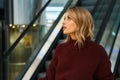 Side view of beautiful blond girl in knitted sweater dreamily looking away on escalator in modern shopping mall Royalty Free Stock Photo