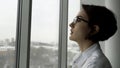 Side view of an elegant woman with short, dark hair and black glasses standing by office window with fuzzy grey city