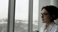 Side view of an elegant woman with short, dark hair and black glasses standing by office window with fuzzy grey city