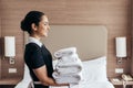 View of smiling maid holding pile of folded towels near bed in hotel room