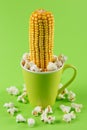 An ear of corn sticks out of a green cup, a symbol of fertility, surrounded by scattered popcorn, concept Royalty Free Stock Photo