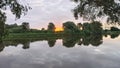 On an early summer morning, the sun rises above the trees on the far side of the river and colors the cloudy sky with delicate ton Royalty Free Stock Photo