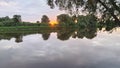 On an early summer morning, the sun rises above the trees on the far side of the river and colors the sky with delicate hues. The Royalty Free Stock Photo