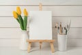 Easel with blank canvas, brushes and tulips on white wooden table Royalty Free Stock Photo