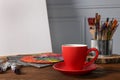 Easel with blank canvas, cup of drink and different art supplies on wooden table near grey wall Royalty Free Stock Photo