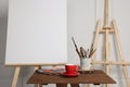 Easel with blank canvas, cup of drink and different art supplies on wooden table near white wall Royalty Free Stock Photo