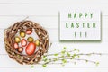 Easter background. HAPPY EASTER written in light box, Easter eggs in willow wreath, birch tree branches on white wooden background
