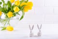 Easter composition with bouquet of yellow tulip flowers in glass vase and two white ceramic rabbits on white Royalty Free Stock Photo