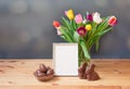 Easter concept. Chocolate Easter eggs in wicker basket, chocolate bunny, tulip flowers and white frame on wooden table Royalty Free Stock Photo