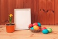 Easter concept. Little daffodil in flower pot, Easter eggs in wicker basket and white frame on wooden table