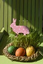 Easter decor with a pink toy rabbit and colorful eggs in a basket. Vertical. Sunlight.