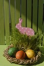 Easter decorations with a toy rabbit and colorful eggs in a basket against a green fence. Vertical. Sunlight.