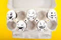 Easter eggs with drawn cartoon faces in a cardboard tray on yellow background.