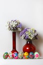 Easter eggs. They are in a row. In the background two red vases of different sizes. In the vases of flowers