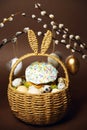 Easter holidays, cute basket with bunny ears on brown background. Easter cakes with colored sprinkles, colored golden eggs, and