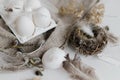 Easter rustic still life. Easter natural eggs in tray, feathers, willow branches, nest on burlap on white wooden table. Simple