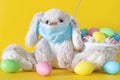 Easter bunny in medical face mask and colored eggs in basket on yellow background. Ester holiday during corona virus concept