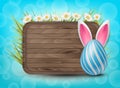 Easter wooden sign with painted 3d realistic eggs with bunny ears in green glass and flowers on bright blue bokeh backdrop.