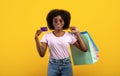 Easy shopping. Excited african american woman holding credit card and shopper bags, posing over yellow studio background