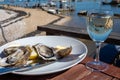 Eating of fresh live oysters with glass of white wine at farm cafe in oyster-farming village, with view on boats and water of