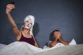 Eccentric housewife with makeup facial mask and towel taking selfie in bed and husband with desperate face expression in weird man