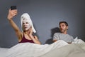 Eccentric housewife with makeup facial mask and towel taking selfie in bed and husband with desperate face expression in weird man