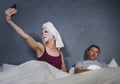 Eccentric housewife with makeup facial mask and towel taking selfie in bed and husband with desperate face expression in weird man