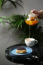 Eclair on ceramic plate anf cup of tea on marble stone and tropic plants on background. tea pot pouring. woman hand hold teapot.