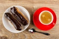 Eclairs with chocolate glaze in plate, cup with coffee on saucer, spoon on plate. Top view