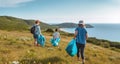 Eco activists of different ages in sportswear with blue bin bags collecting trash on green grassy hillside near calm blue sea on Royalty Free Stock Photo