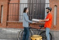 Ecological city, bike and delivering service. Smiling courier in helmet gives pizza to client, near entrance, at street Royalty Free Stock Photo