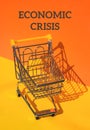 ECONOMIC CRISIS text against Empty shopping trolley cart on isometric orange yellow background. Small toy supermarket